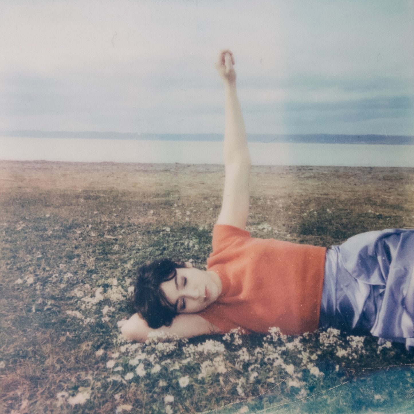 Person lying in a field with arm raised, cloudy sky backdrop.