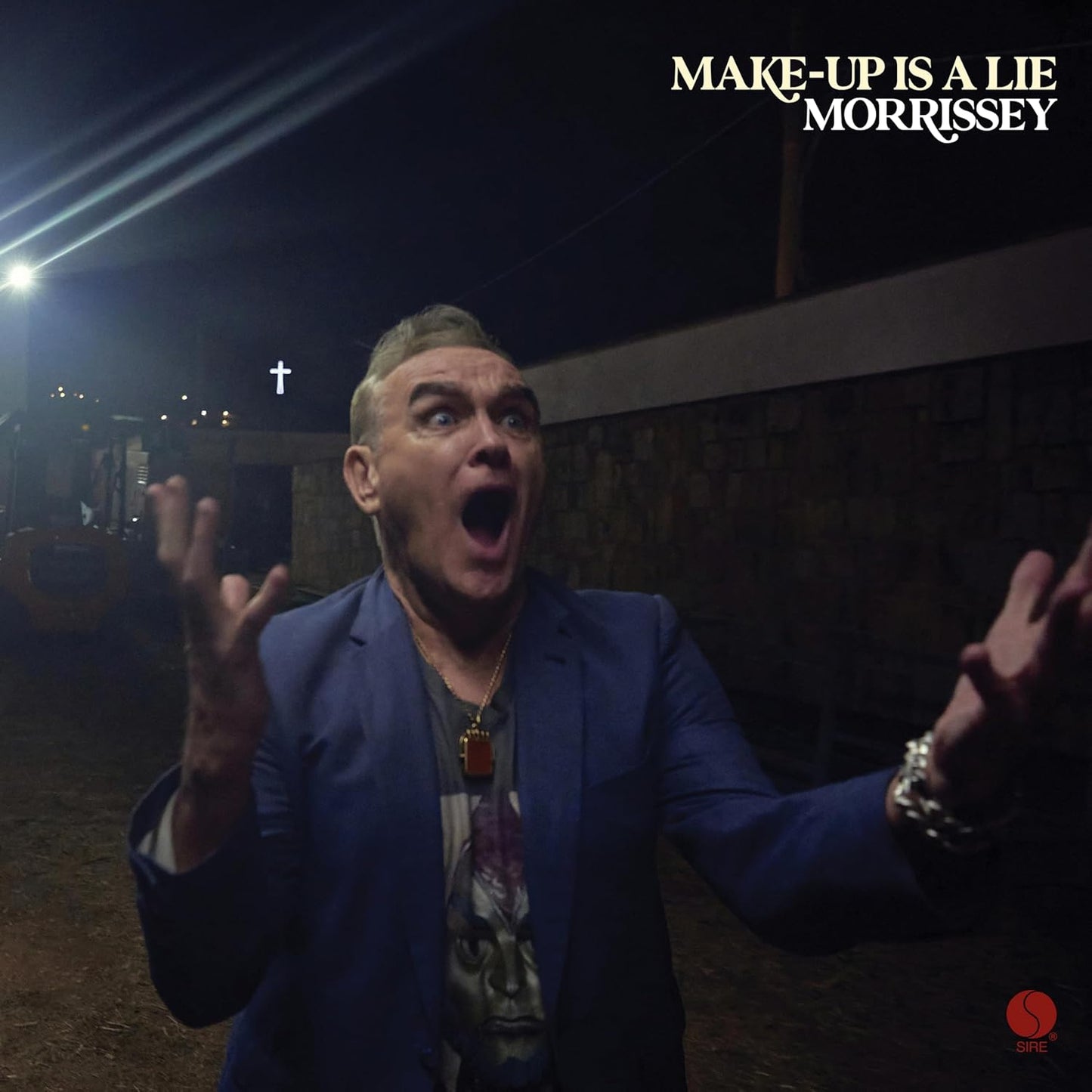 Morrissey with a shocked expression, hands raised, wearing a blue blazer at night.