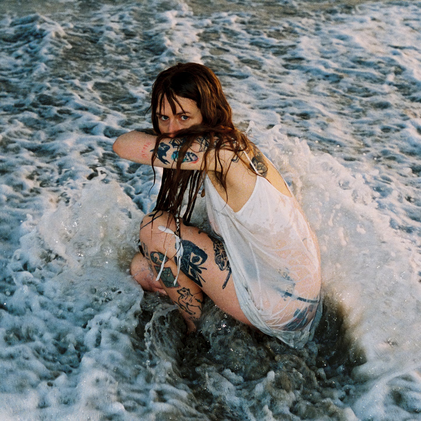 Woman with tattoos kneels in foamy ocean waves, looking at viewer.