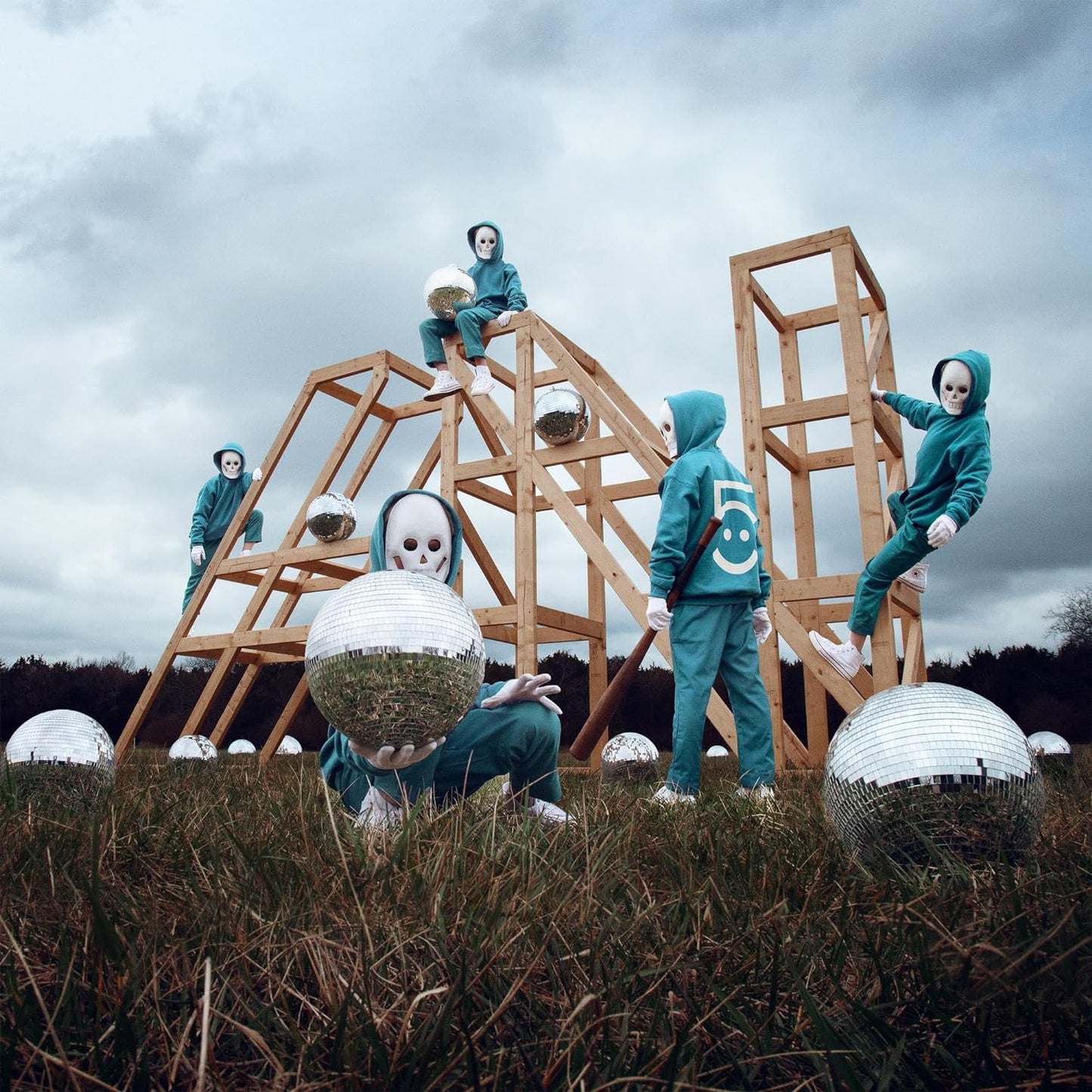 Five figures with skull masks pose amongst disco balls and wooden structures.
