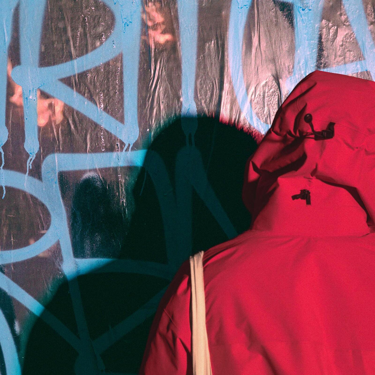 Red hooded figure stands before a wall covered in blue graffiti.