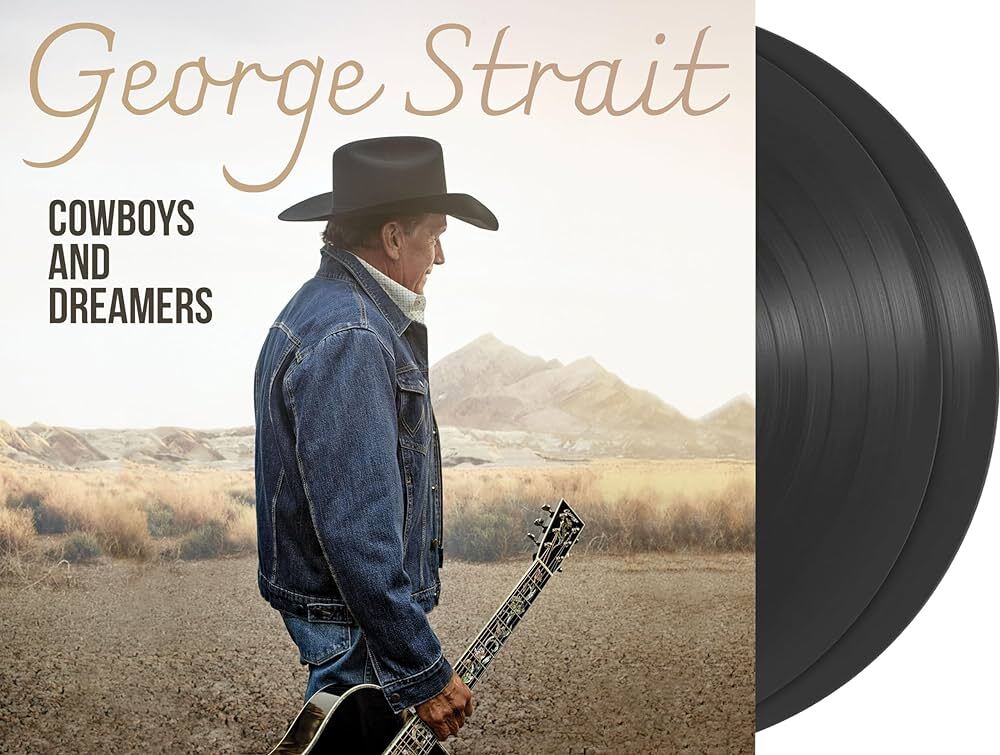 George Strait in a cowboy hat and denim jacket holding a guitar in desert landscape.