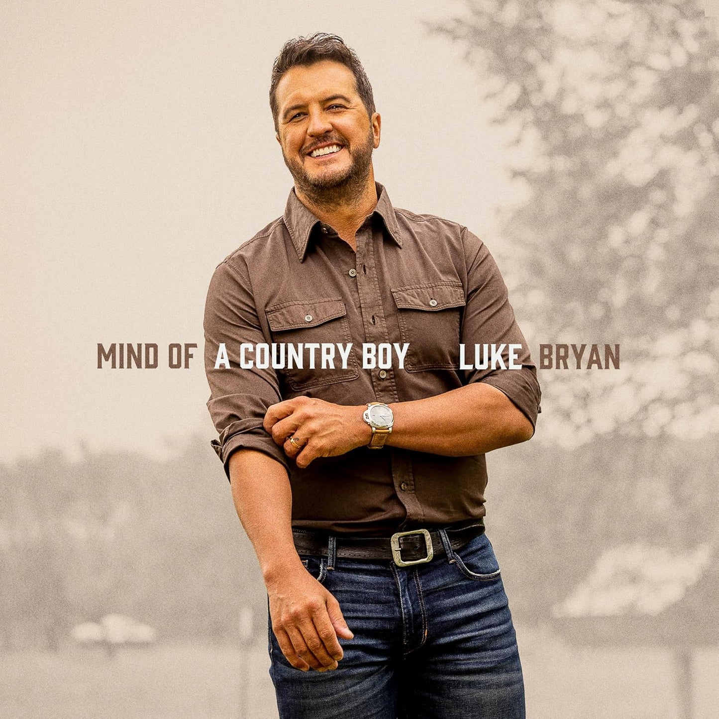 Luke Bryan smiling, wearing a brown button-down shirt and jeans against a blurred background.
