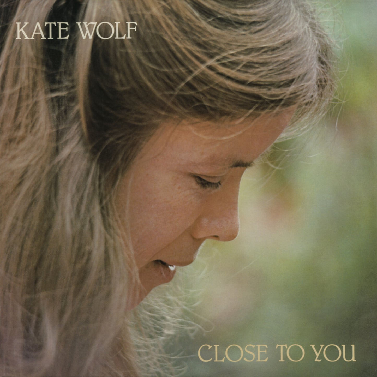 Profile portrait of Kate Wolf with long hair against a blurred outdoor background.