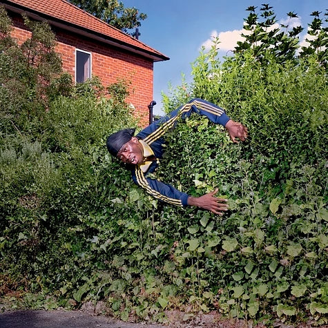 A man wearing a durag and track jacket peeking out from a dense green hedge.