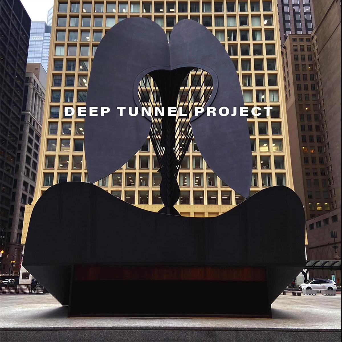 Black metal sculpture with “Deep Tunnel Project” text, city buildings backdrop.