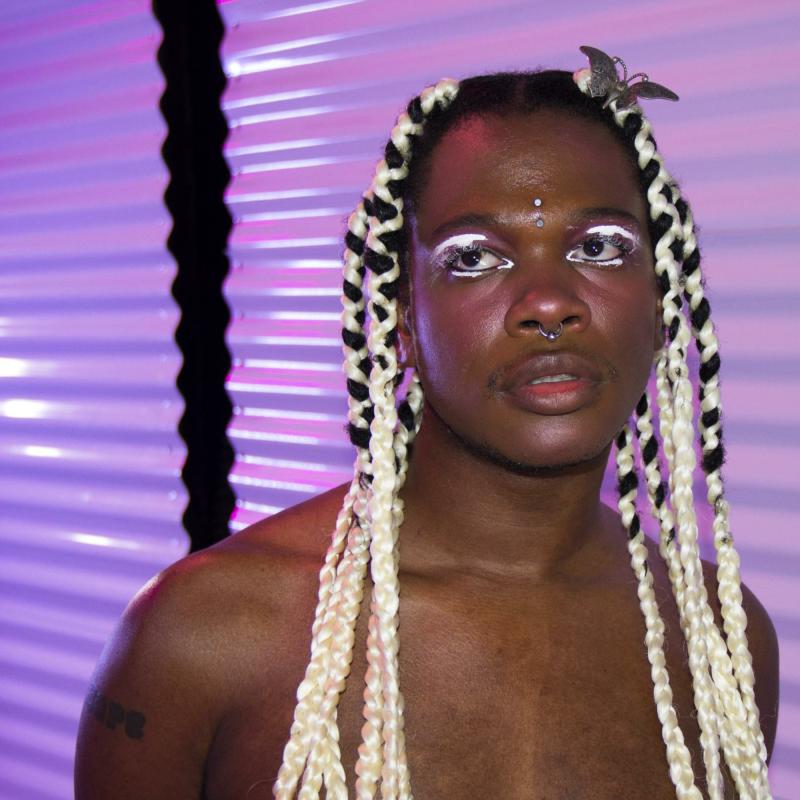 Portrait of a person with blonde braids, white makeup, and a nose ring.