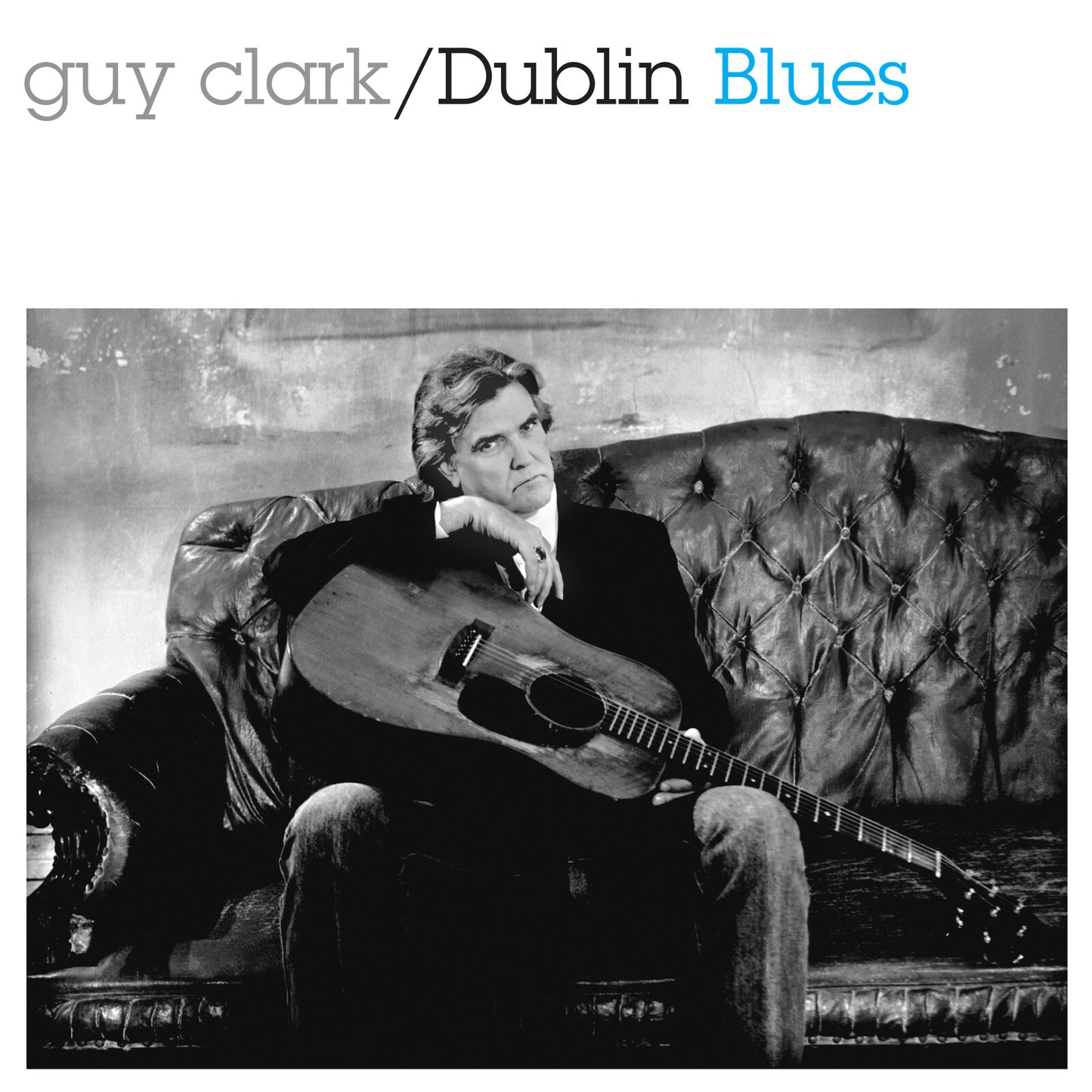 Guy Clark sits on a tufted leather sofa holding an acoustic guitar.