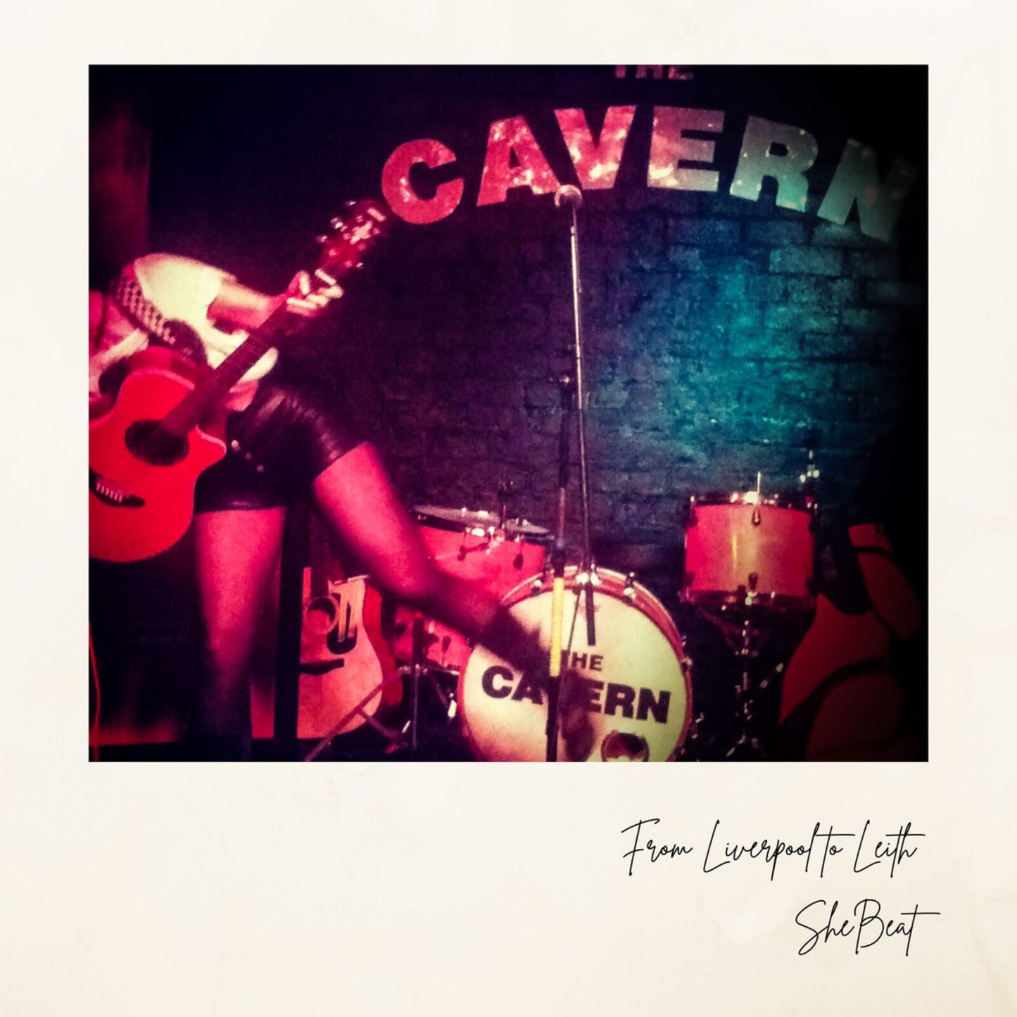 Musician playing guitar in front of a drum kit with "CAVERN" sign.