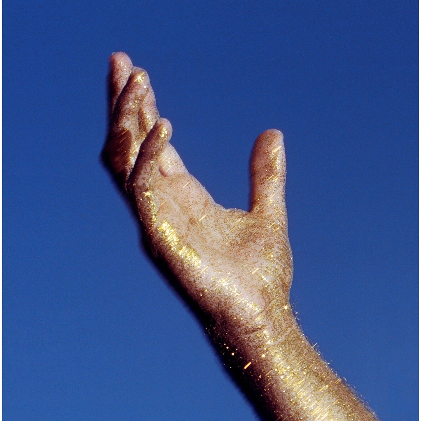 Hand covered in gold glitter reaching upwards against a blue background.