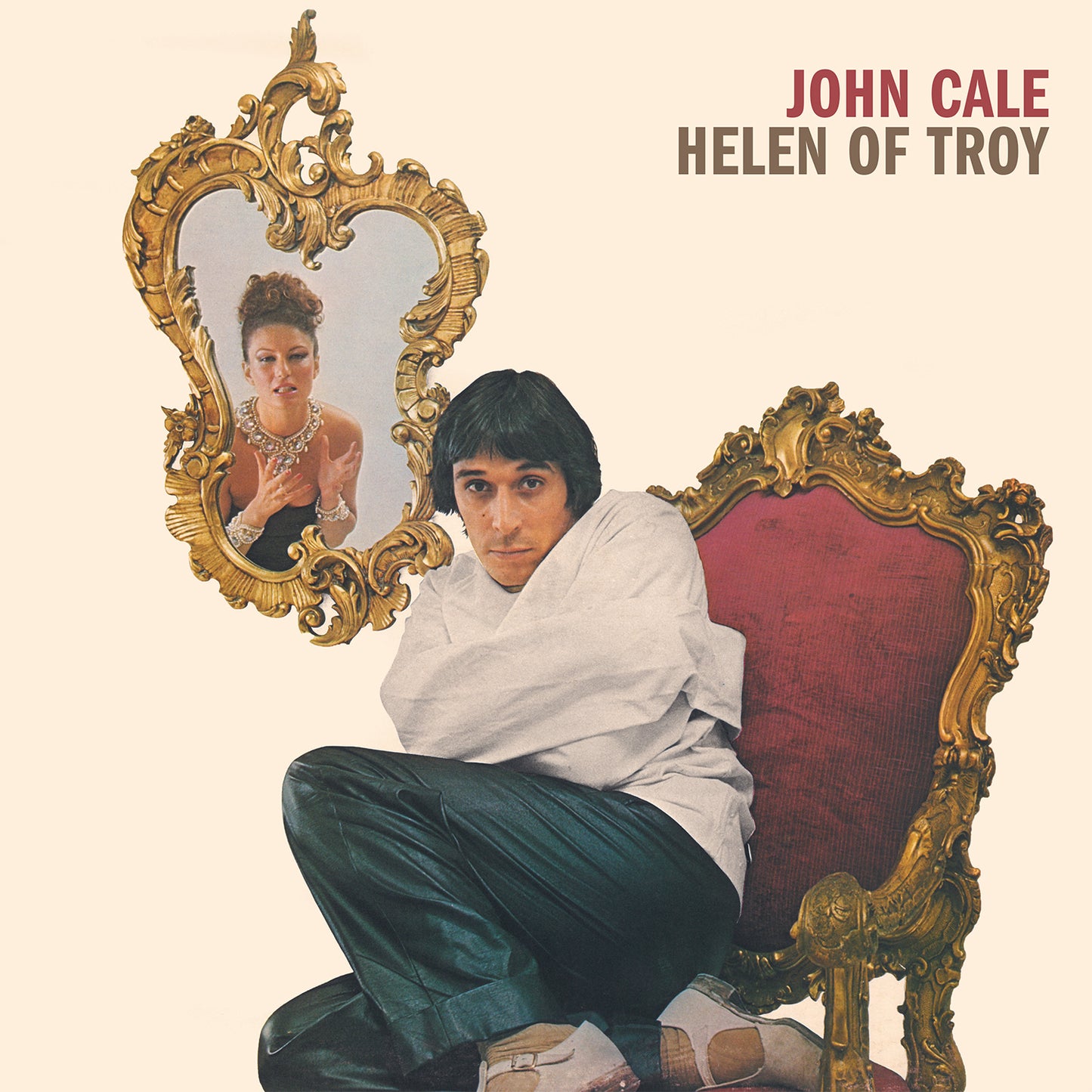 John Cale in a straightjacket sits in a chair next to an ornate mirror.