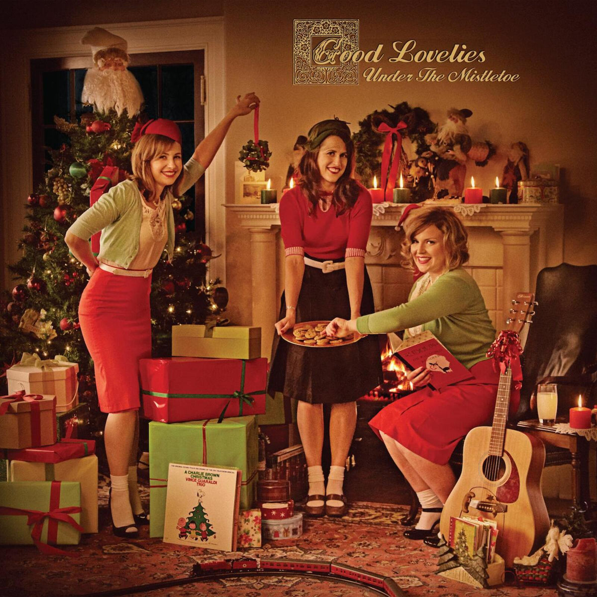 Three women in festive attire pose by a Christmas tree and gifts.