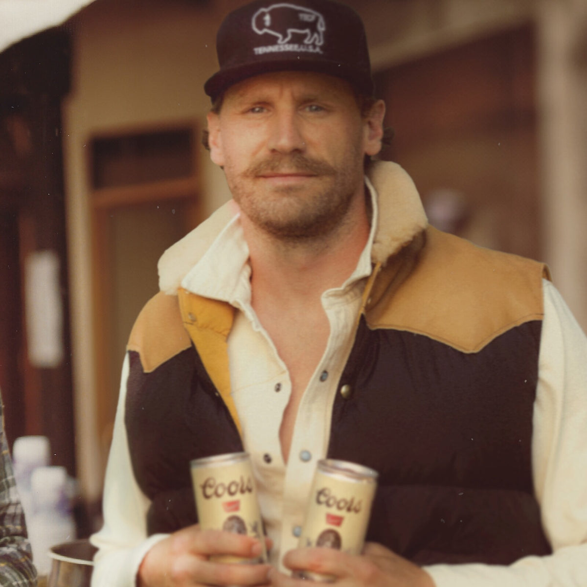 Chase Rice portrait, holding two Coors beer cans, wearing a cap and vest.