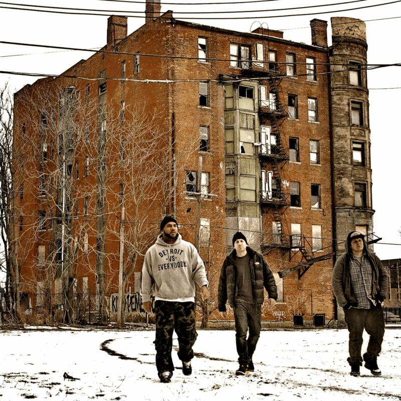 Three men walk towards the camera in front of a brick building.