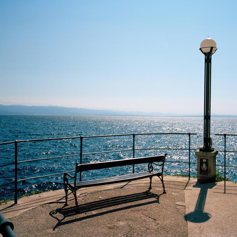 Seaside bench and lamppost with a shimmering ocean and distant hills.