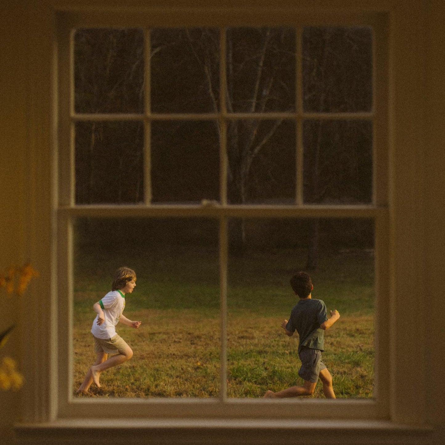 Two children run through a grassy field, viewed through a window frame.