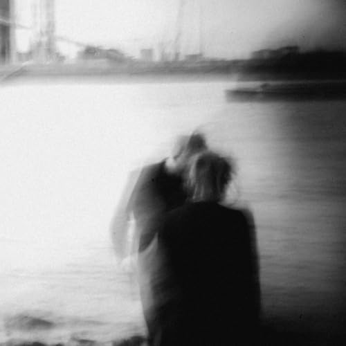 Blurred black and white photo of two figures by water and a bridge.