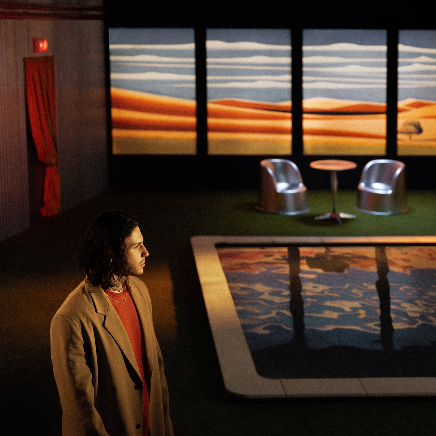 Man with long hair stands beside a pool with desert backdrop.