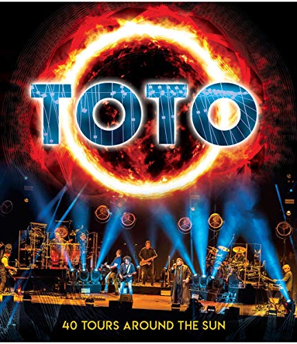 Toto performing on stage under a large glowing sun graphic with album title text.