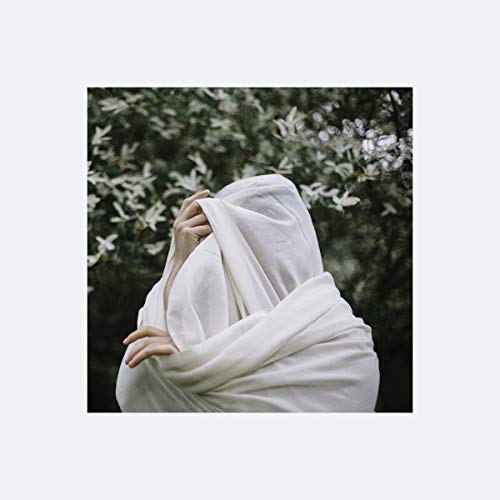 Person wrapped in white fabric, facing away, with blurred foliage background.