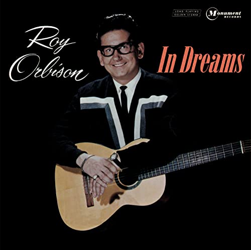 Roy Orbison wearing glasses and a suit, holding an acoustic guitar against black background.