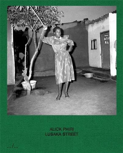 Black and white photo of woman in dress standing beside a small tree outdoors.