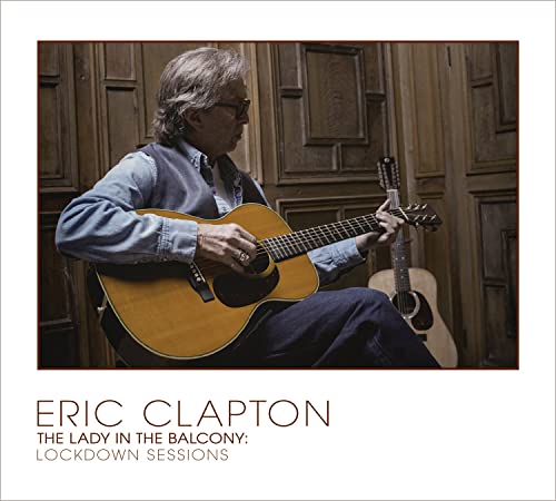Eric Clapton playing an acoustic guitar, seated indoors with wood paneling.