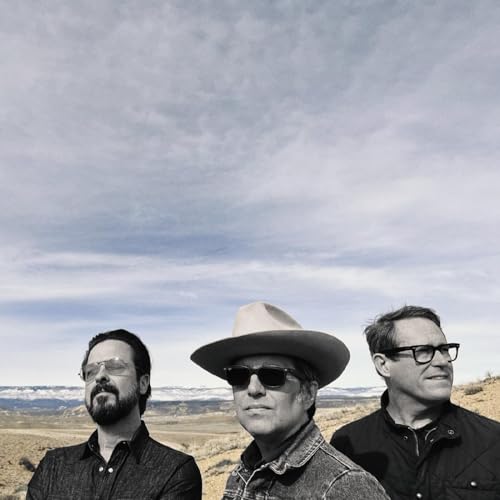 Three men in sunglasses and hats stand before a desert landscape.