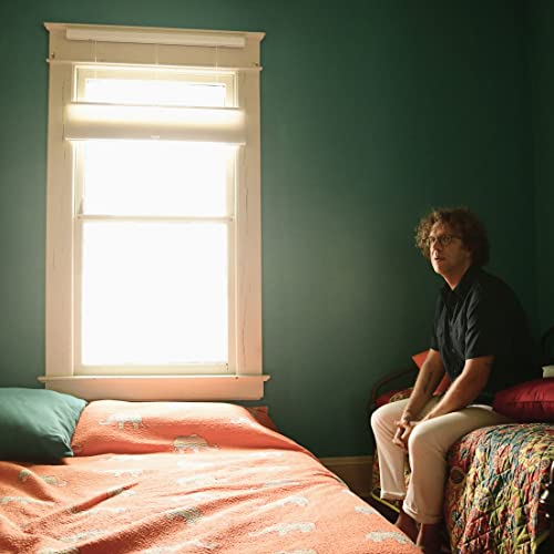 A man with curly hair sits on a bed near a window in a room.