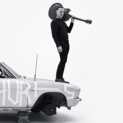Black and white photo of Bryan Adams standing on a car with a guitar.
