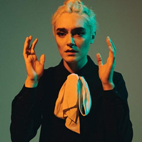 Portrait of a pale woman with short blonde hair, hands raised.