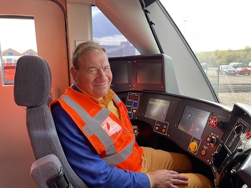 Michael Portillo sitting in a train driver's cab wearing a high-visibility vest.