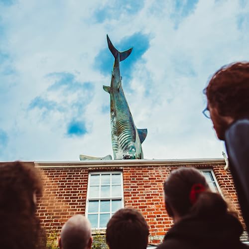 Large shark sculpture atop a brick building, viewed from a crowd.