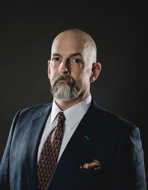A bald man with a gray goatee wearing a dark suit and patterned tie.