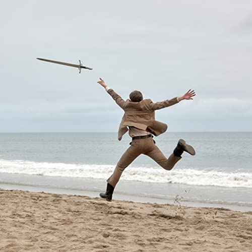 Man in suit leaps with sword against a beach and sky.