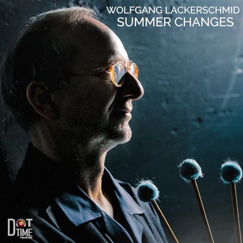 Profile portrait of Wolfgang Lackerschmid wearing glasses, holding three mallets against a dark background.