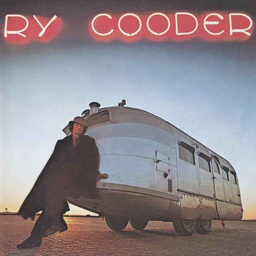 Ry Cooder leaning against a silver vintage trailer on a desert landscape at sunset.