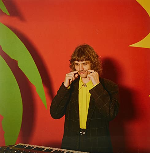 A man with curly hair wearing a plaid blazer and yellow shirt smiles.