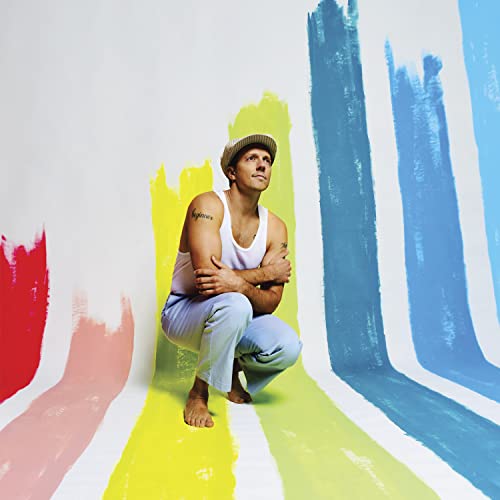 Jason Mraz kneels against a colorful, striped wall, looking upwards.
