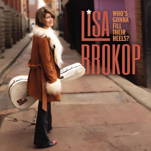 Lisa Brokop in a fur-trimmed coat carries a guitar case down an alleyway.