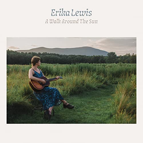 Woman with guitar sits in a field with mountains in the distance.