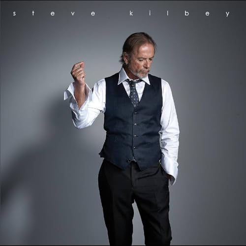Steve Kilbey portrait, wearing a waistcoat and tie, grey background.
