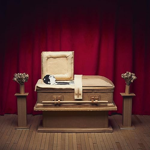 Man lying in a cardboard coffin against a red curtain backdrop.