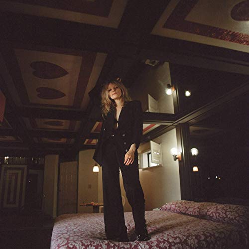 Jessica Pratt stands in a room with patterned walls and a bed.