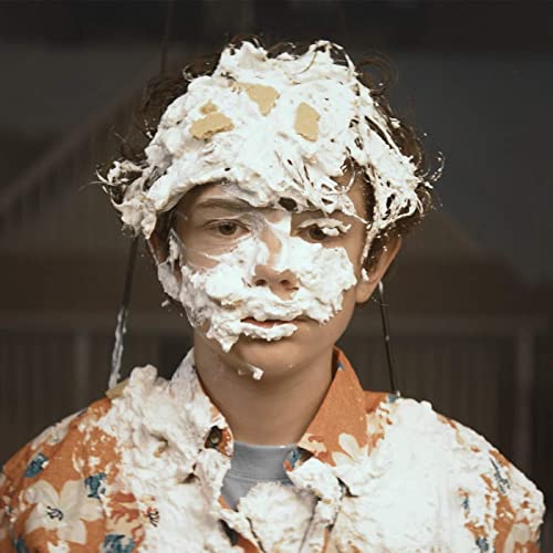 Person covered in white cream, looking directly at the viewer.