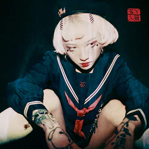 Pale-haired person in a dark sailor suit, with facial piercings and tattoos.