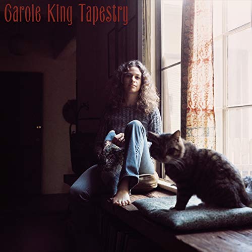 Carole King sits barefoot on a windowsill with a cat beside her.