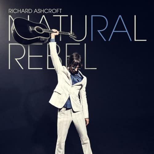 Richard Ashcroft in a light suit holding an acoustic guitar above his head.