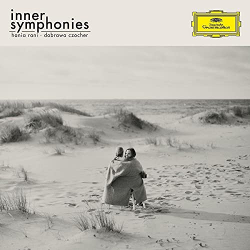 Two people wrapped in a blanket sit on a sandy beach in black and white.