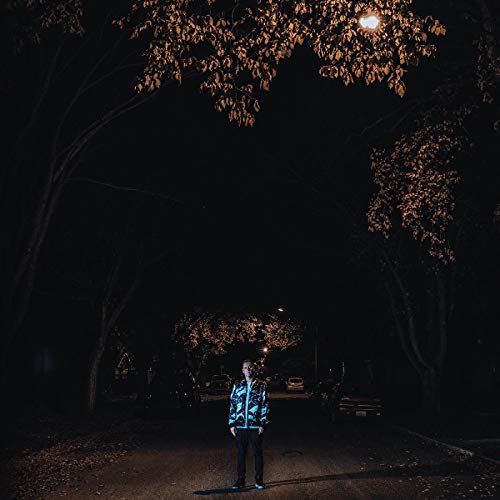 Person in patterned jacket standing on a dark, tree-lined street.
